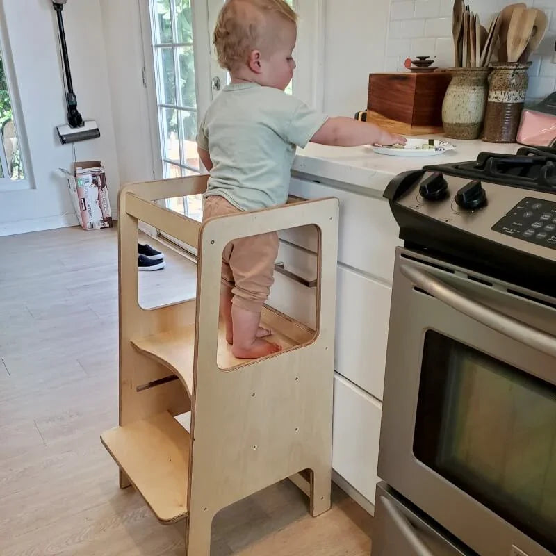 Toddler standing on a Montessori learning tower at a kitchen counter.
