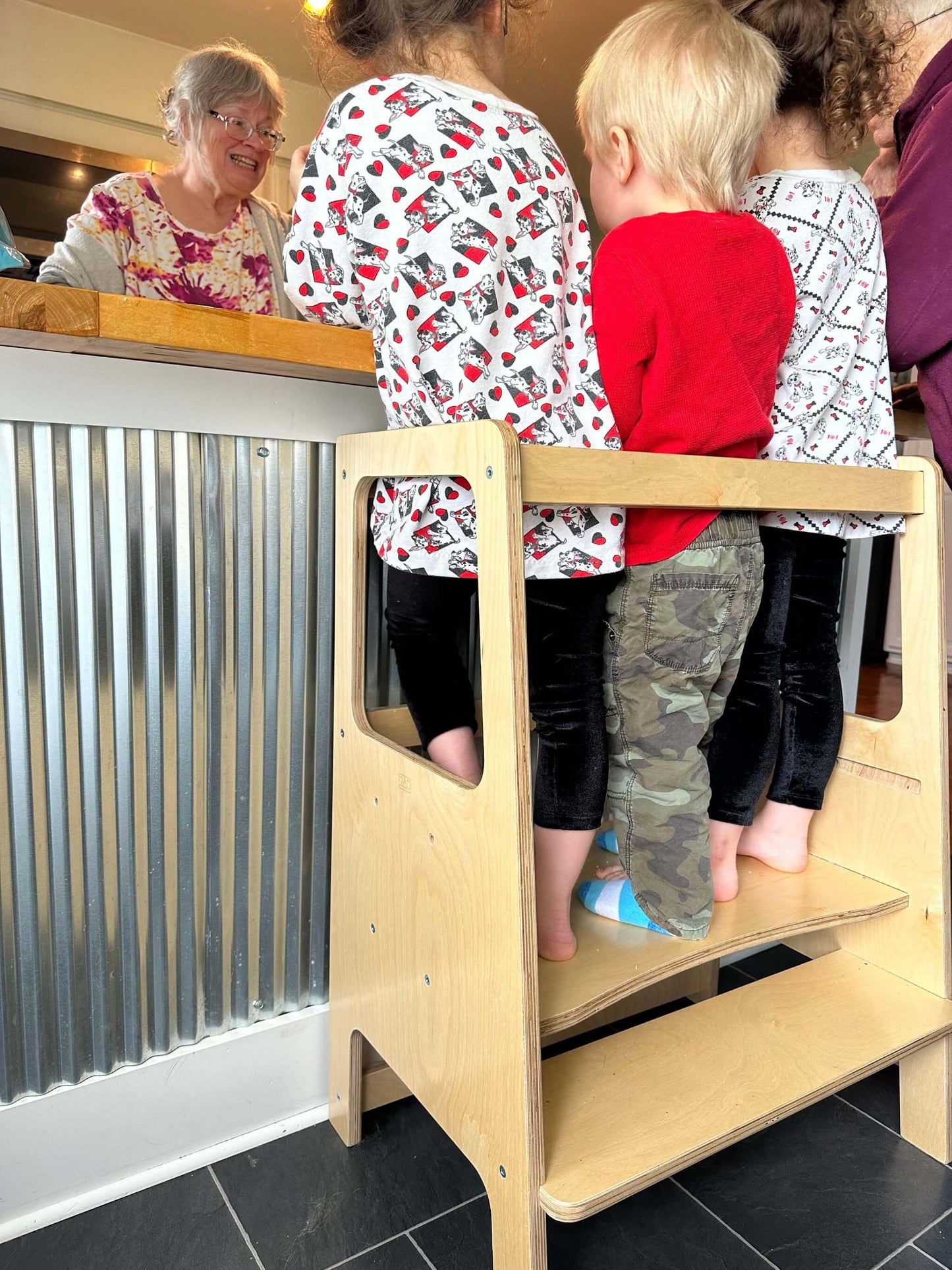 Three young children standing together on a wooden learning tower at a kitchen counter.
