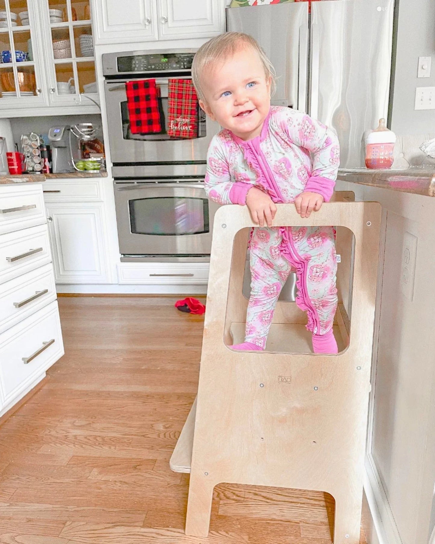 Smiling toddler in pink pajamas stands on a wooden learning tower in a bright kitchen