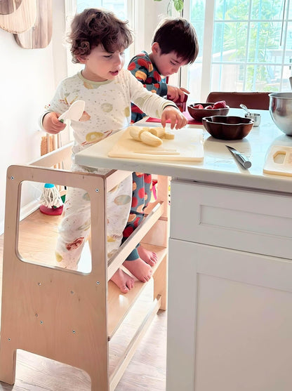 Two young children on a wooden learning tower preparing food at a kitchen counter