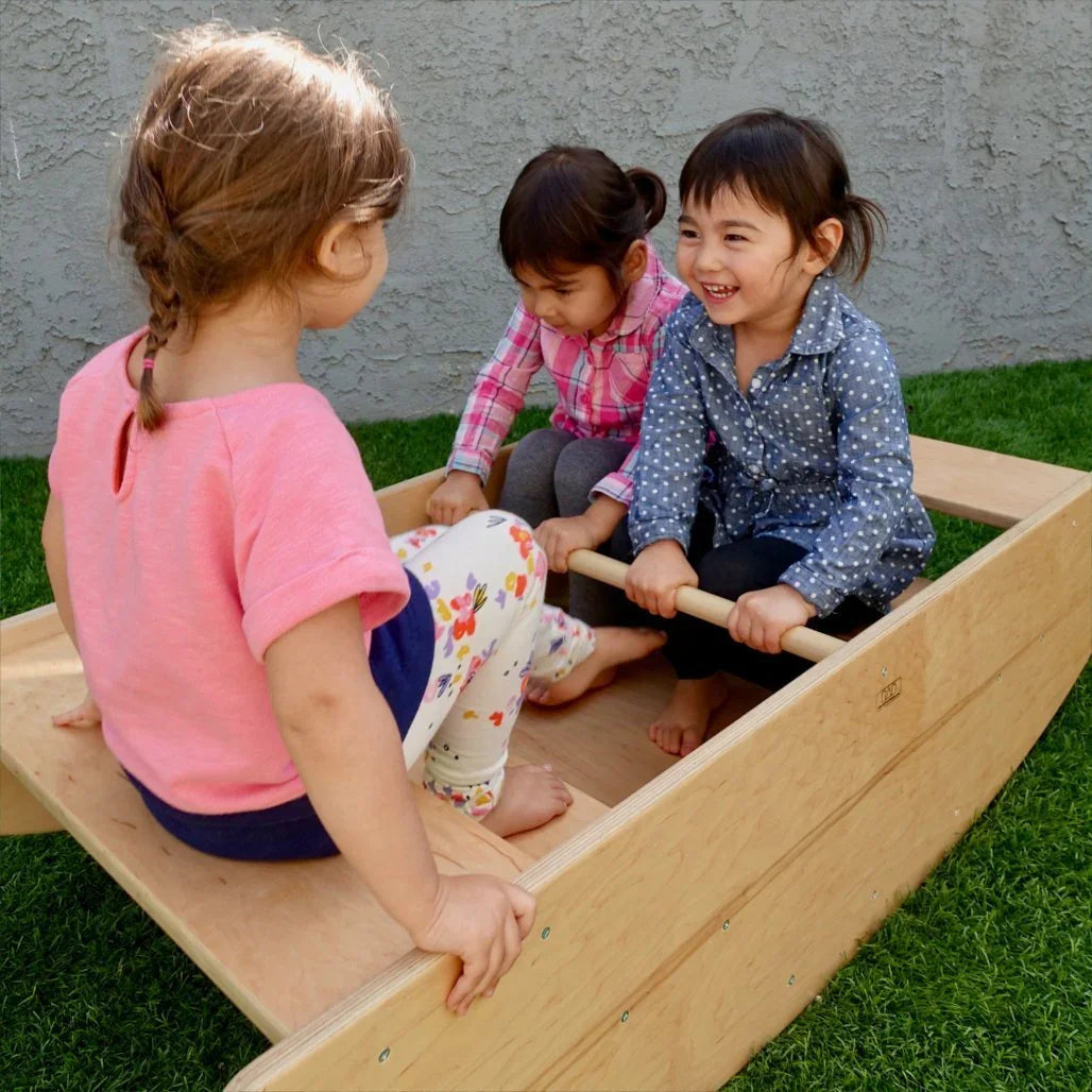 Three young children playing on a wooden sensory rocking boat outdoors on grass