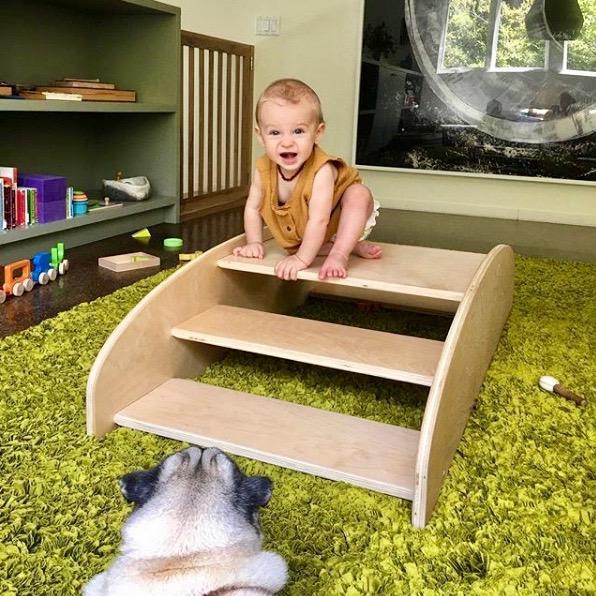 Baby climbing wooden sensory steps on green rug with dog nearby in playroom