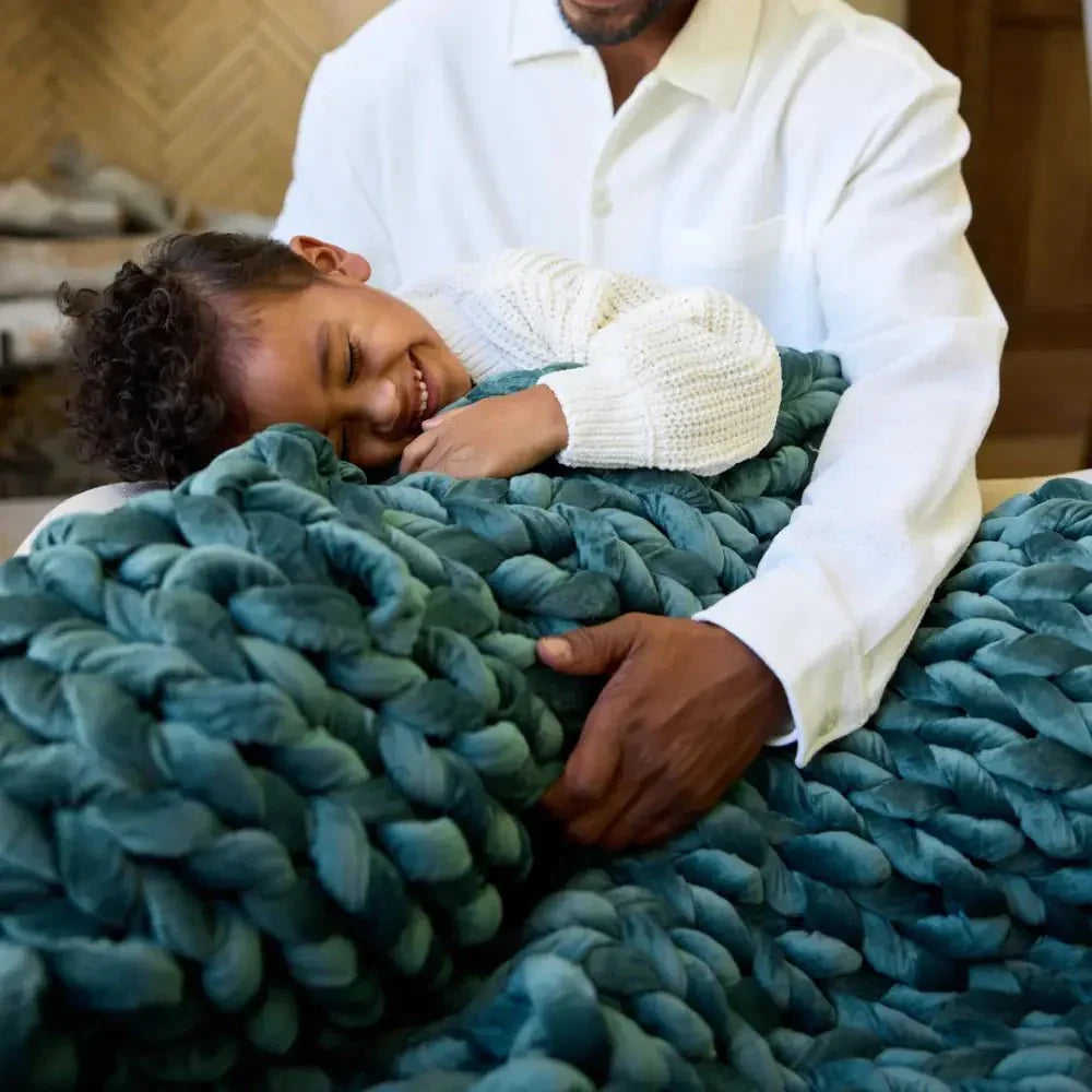Child smiling under chunky sapphire blue weighted blanket with adult, cozy sensory support