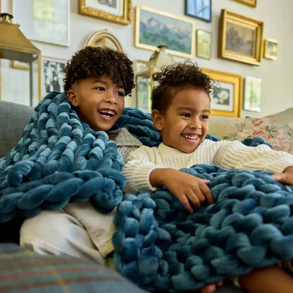 Two smiling children under a chunky sapphire blue velvet weighted blanket on a sofa